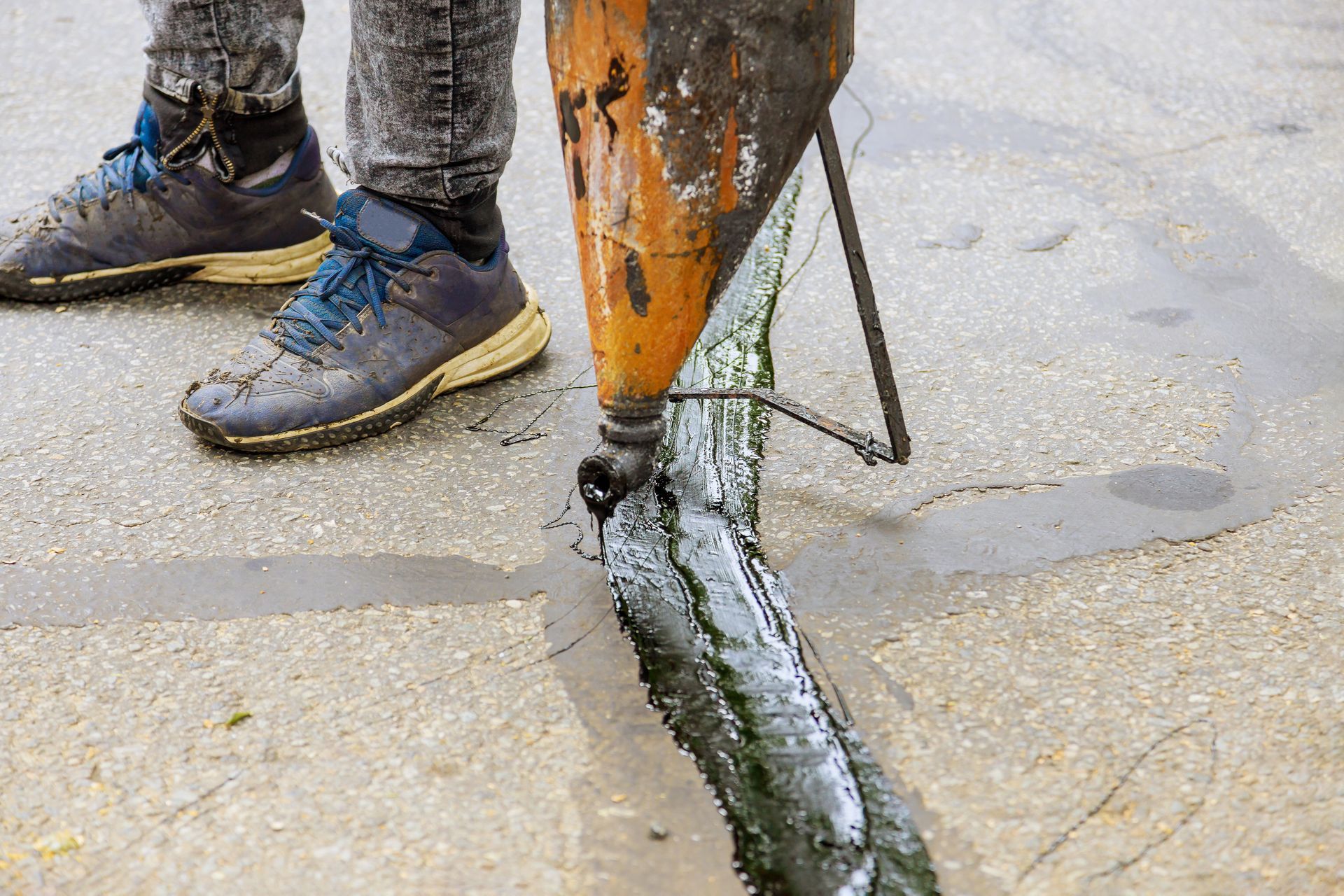 Worker’s muddy boots beside an orange drill cutting into wet pavement.