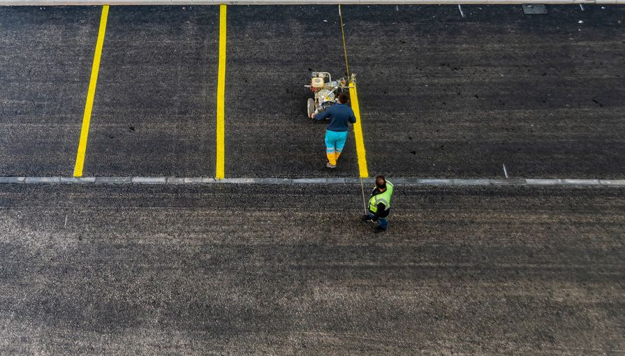 Two people on a track-like surface with yellow lane lines, one carrying gear and one in neon green running nearby