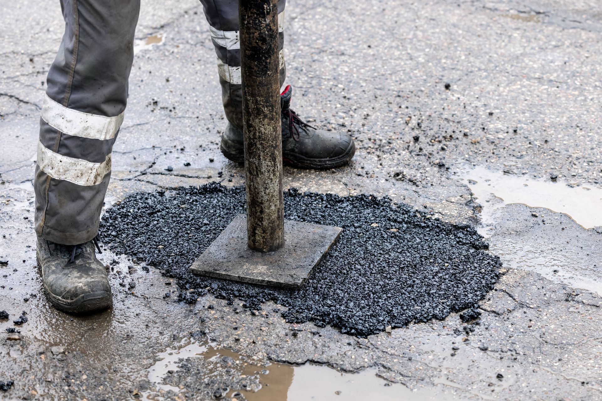 Worker tamping fresh asphalt around a utility pole on a wet street