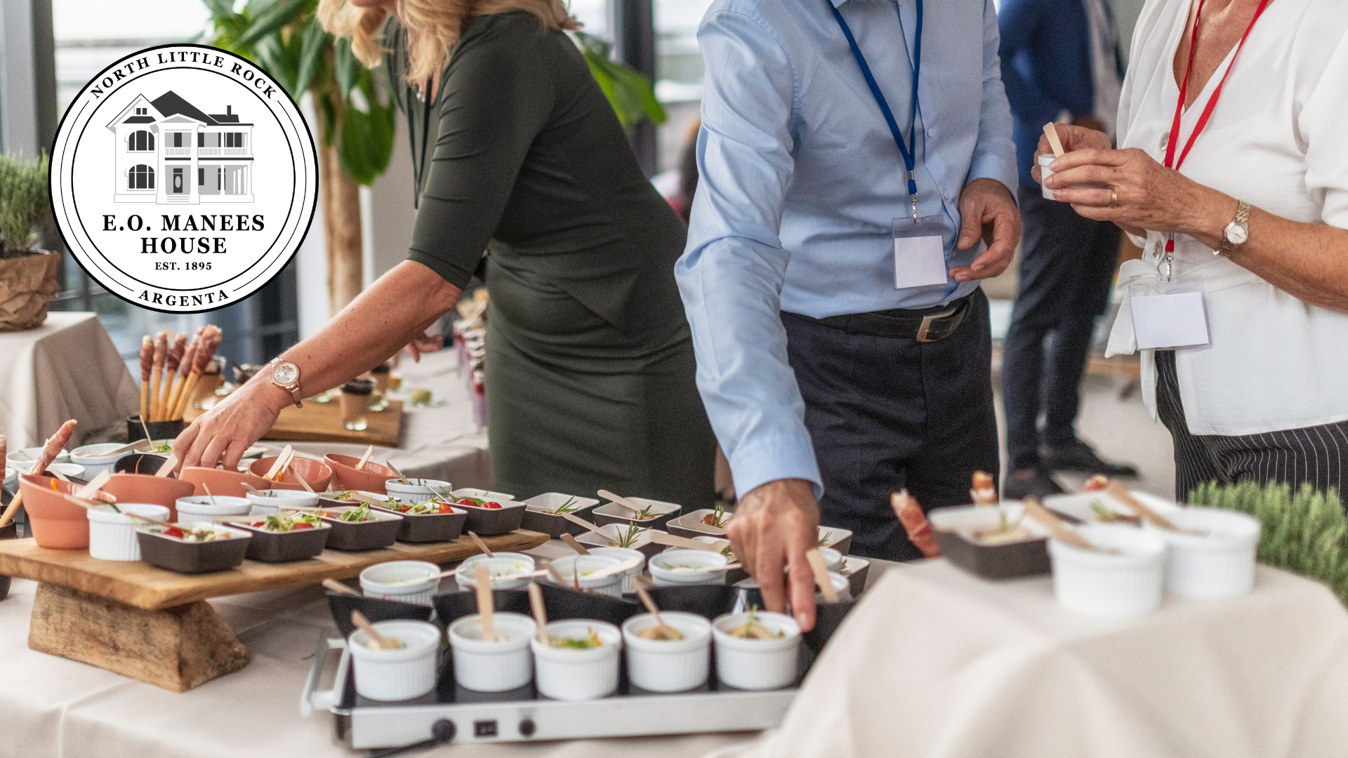 People interact with a catered appetizer buffet at an indoor event, with a circular business logo in the corner.