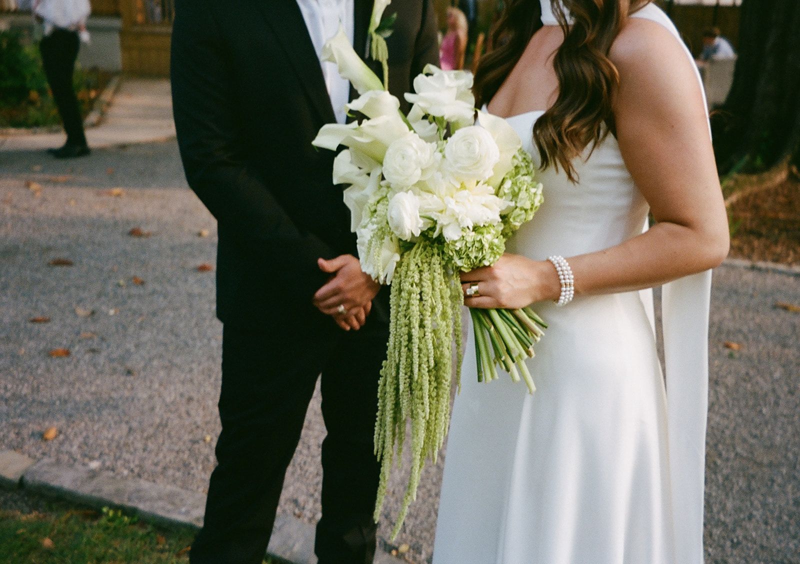A person in a white gown holds a bouquet of white flowers with cascading green stems, standing next to another person.