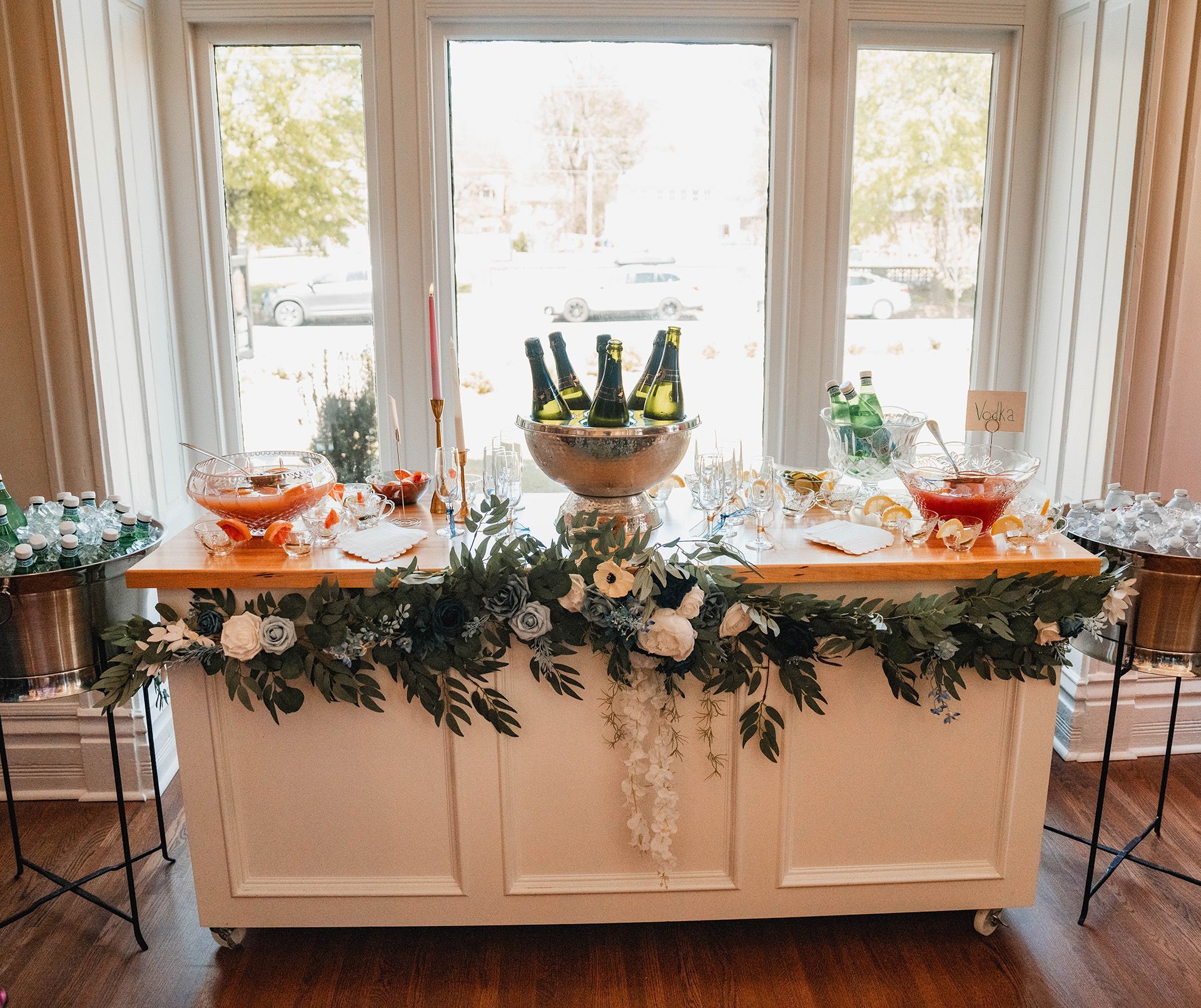 A long white table with flowers and wine bottles on it in front of a window.