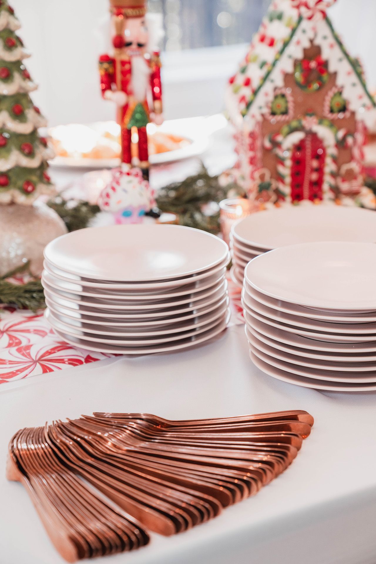 A stack of plates and forks on a table with a gingerbread house in the background.