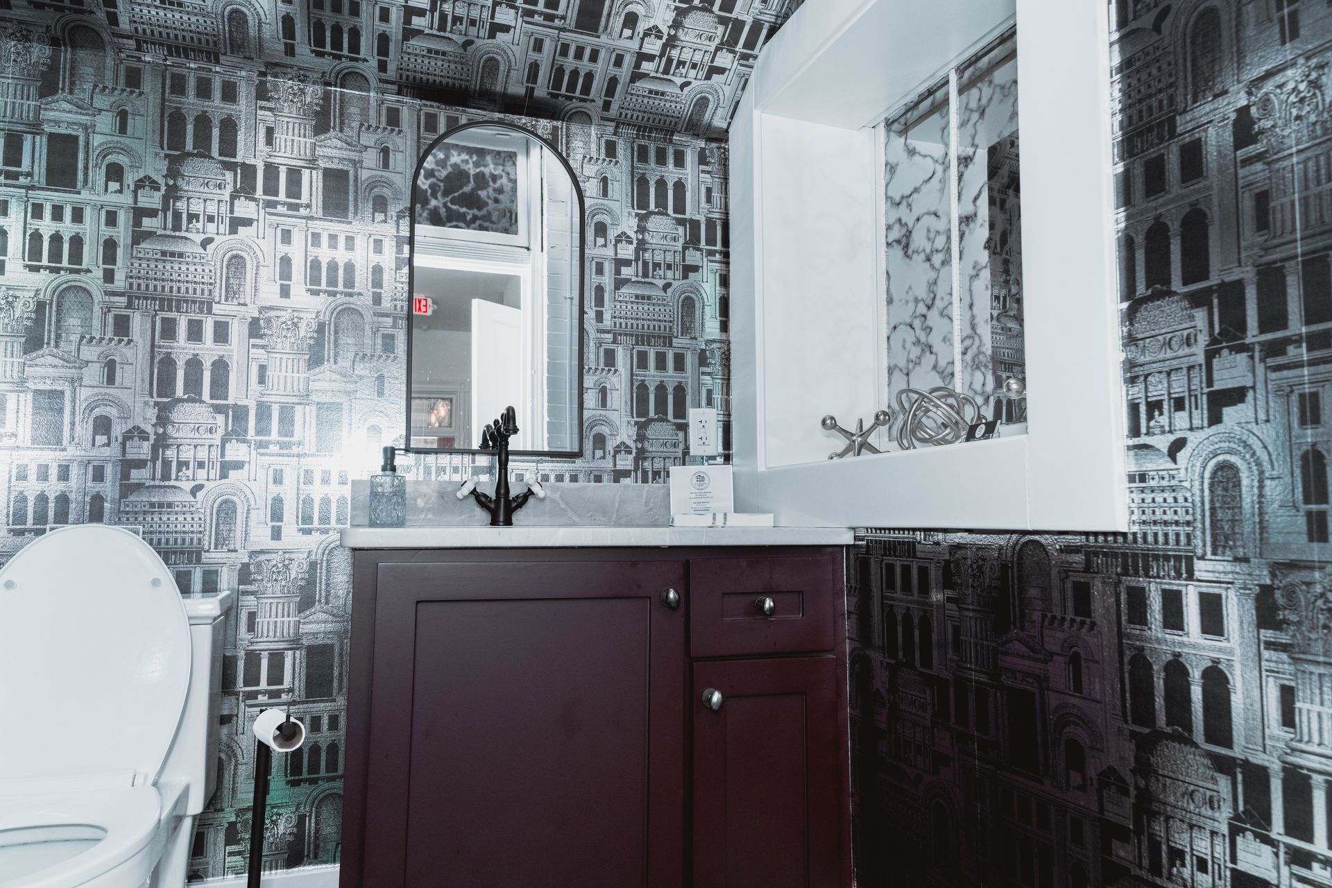 A bathroom with a toilet , sink , mirror and wallpaper.