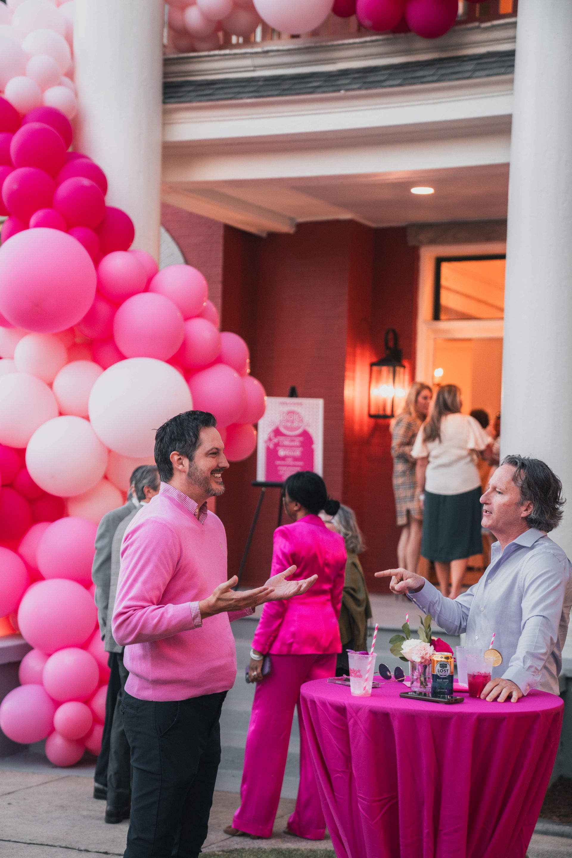 A group of people are standing around a table with pink balloons in the background.