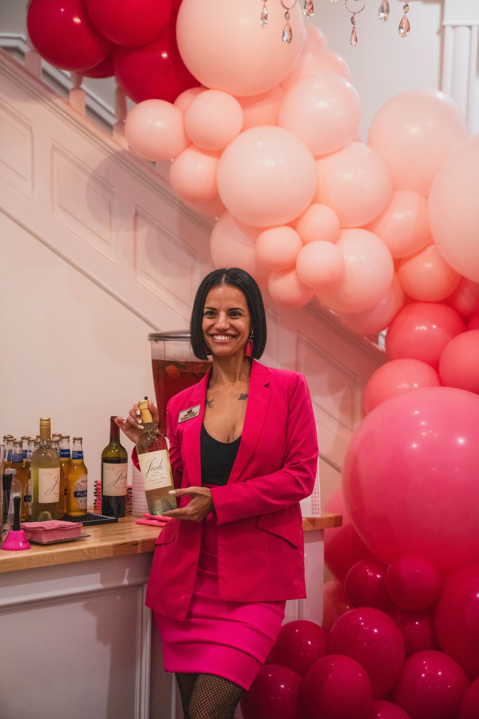 A woman in a pink suit is standing in front of a wall of pink balloons.