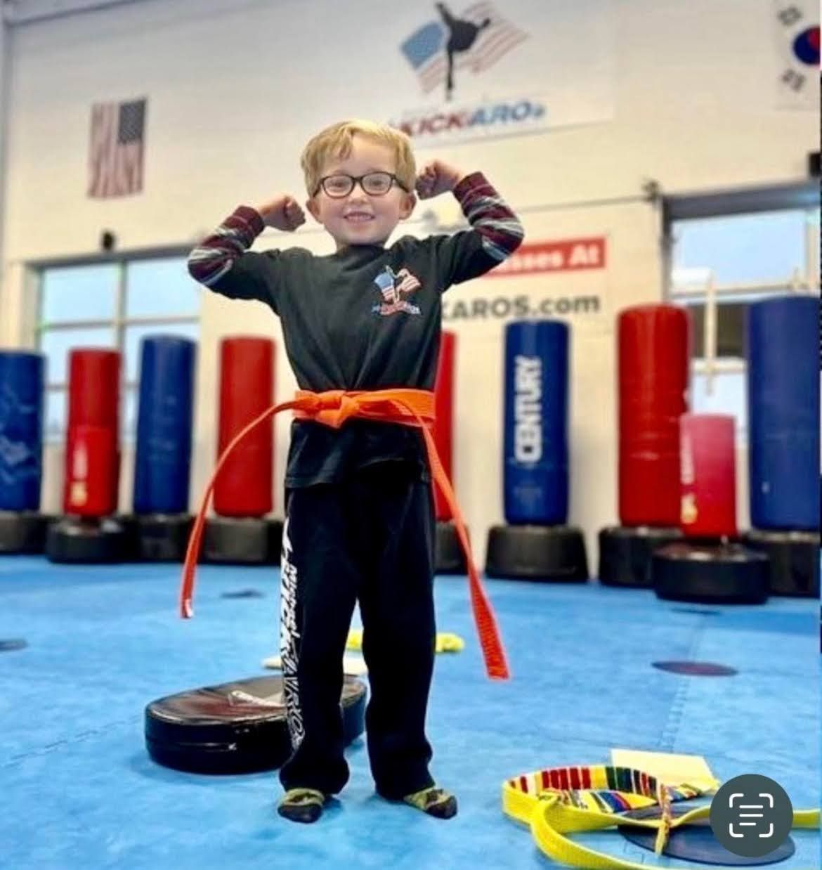 A young boy with an orange belt stands in a gym