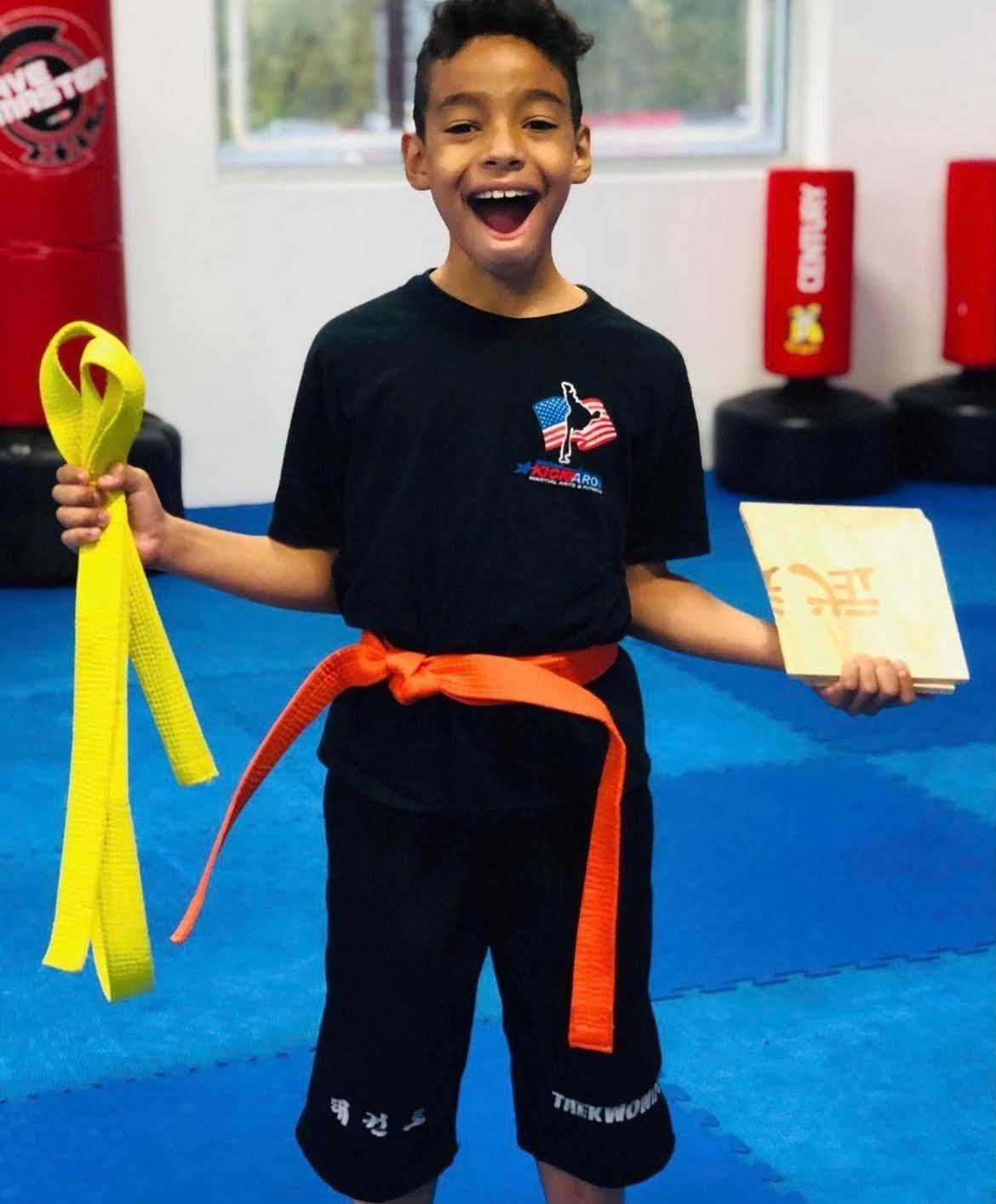 A young boy with an orange belt is holding a yellow belt and a book