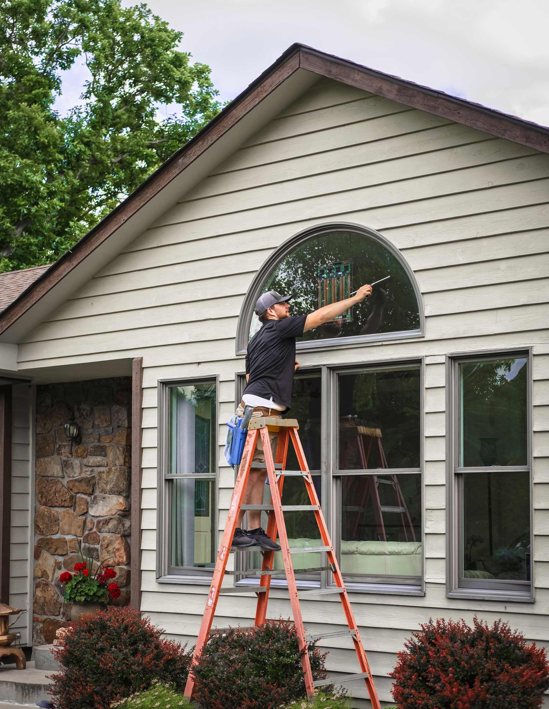 A man is standing on a ladder cleaning a window of a house.