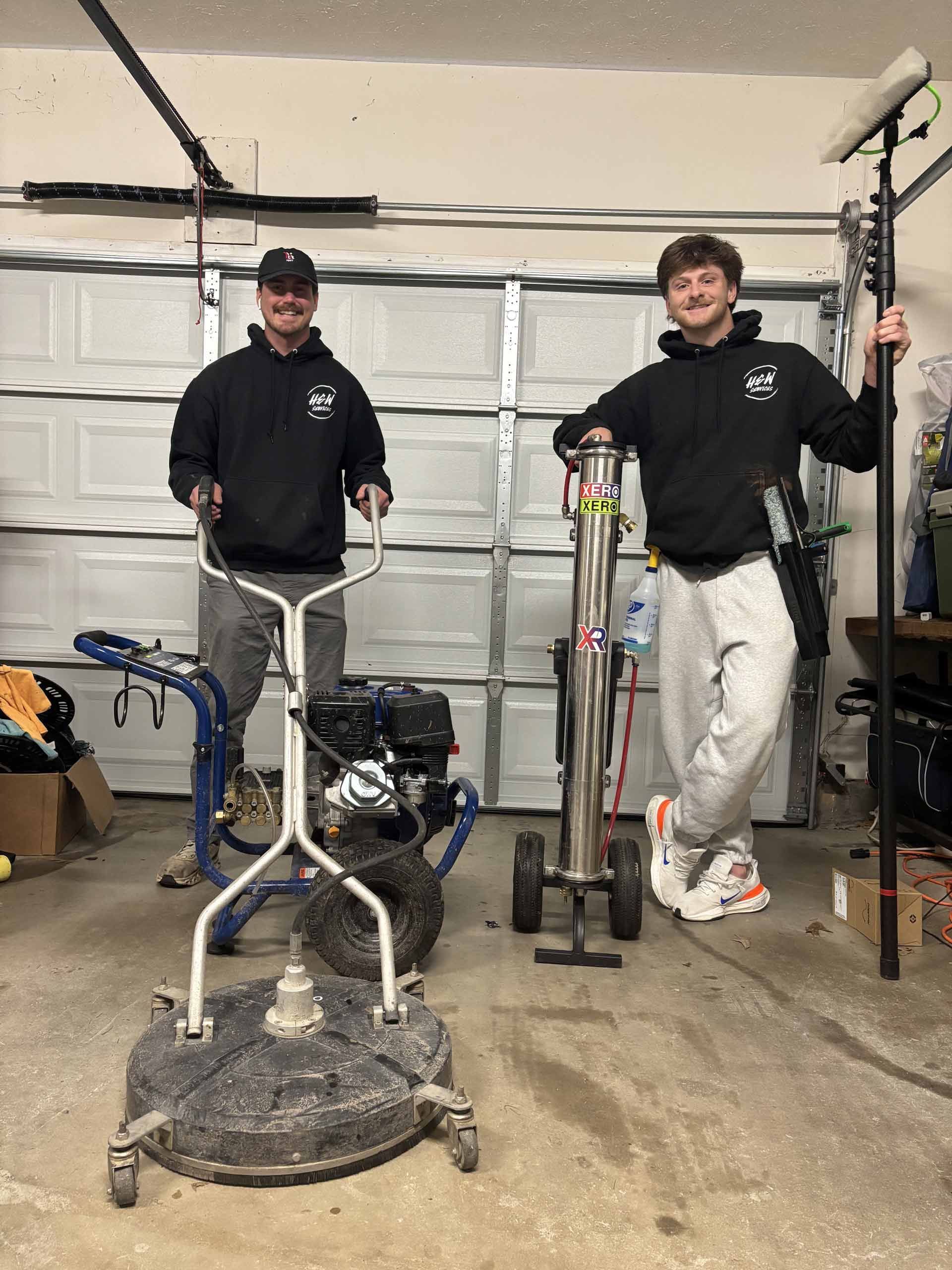 Two men are standing next to each other in a garage holding cleaning equipment.