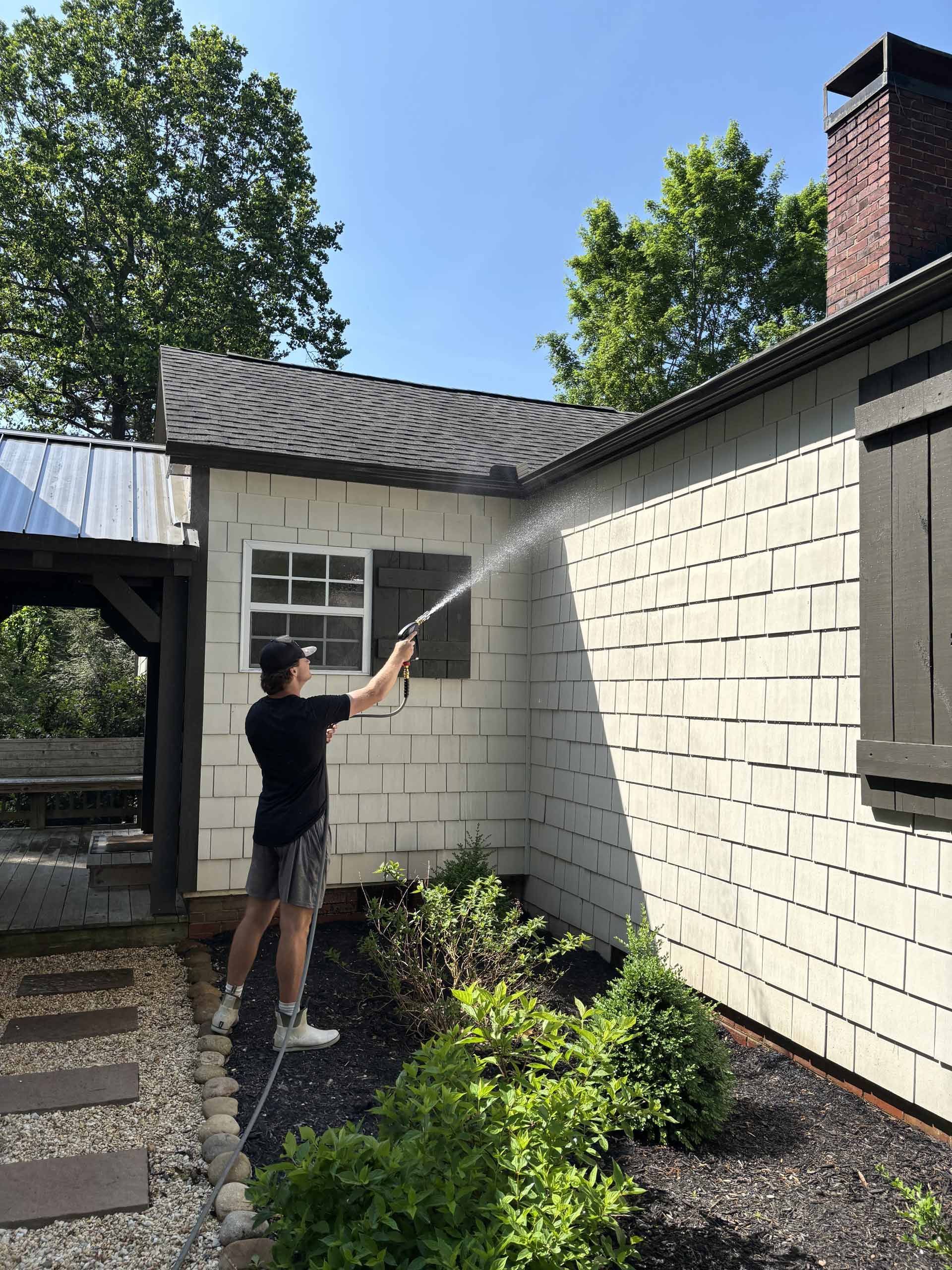 A man is spraying water on the side of a white brick house.