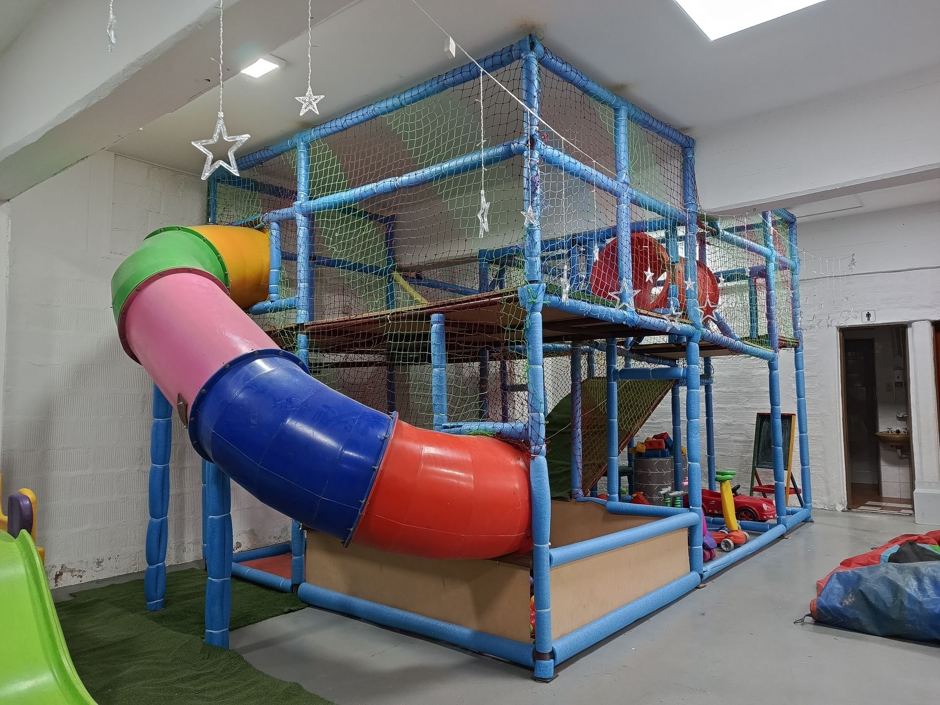 Parque infantil interior con tobogán de tubos de colores, estructura de escalada y red.