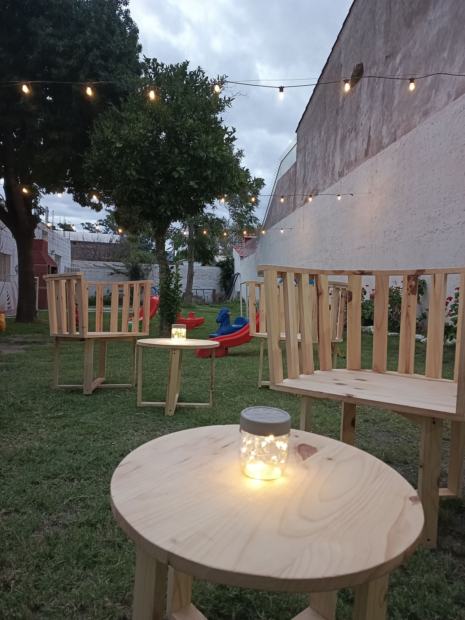 Zona de estar al aire libre con mesas de madera, sillas y luces de cadena, ubicada sobre césped.