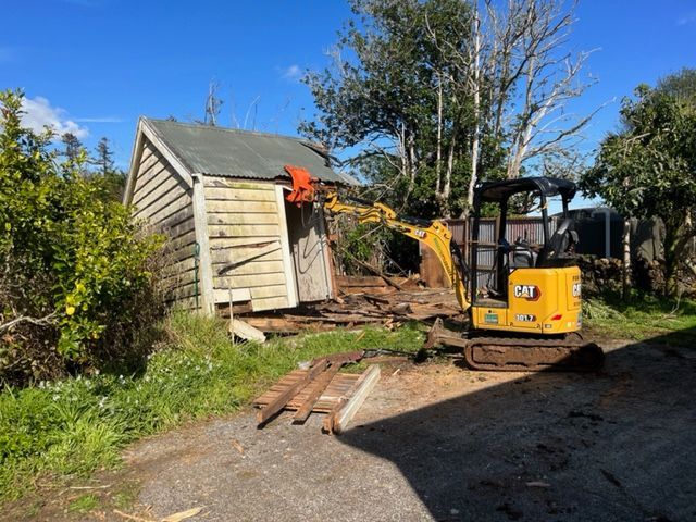 A small yellow excavator is demolishing a shed.