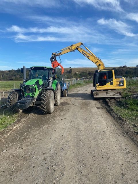 A green tractor is being towed by a yellow excavator on a dirt road