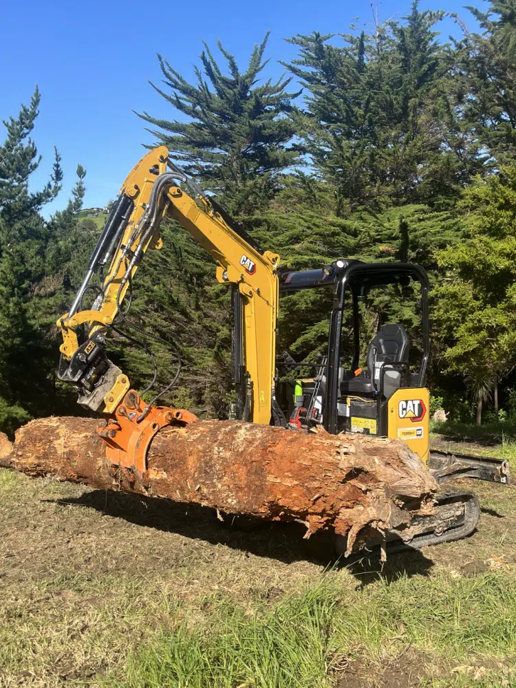 A yellow excavator is carrying a large log in a field.