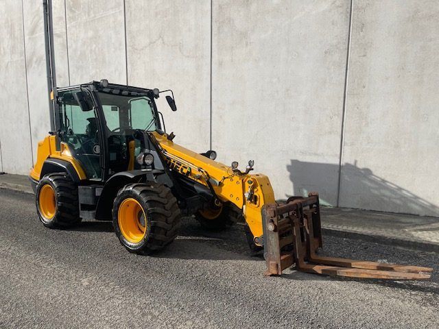A yellow tractor with a forklift attached to it is parked in front of a concrete wall.