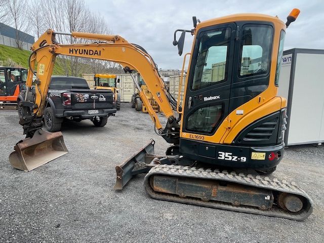 A hyundai excavator is parked in a gravel lot next to a truck.