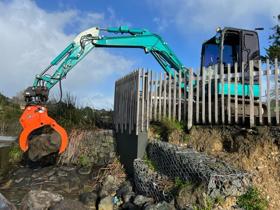 A crane is lifting a large rock in front of a wooden fence.