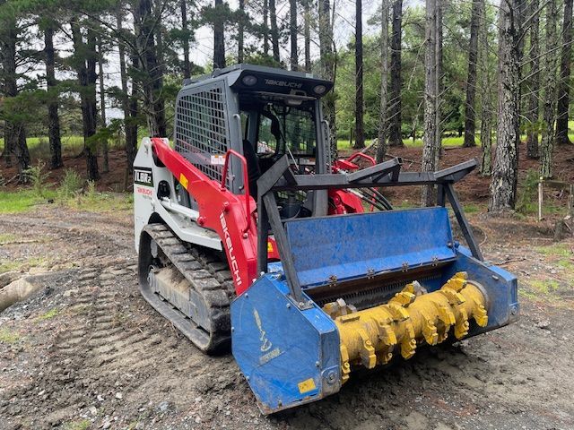 A bulldozer with a blue bucket is parked in the dirt in the woods.