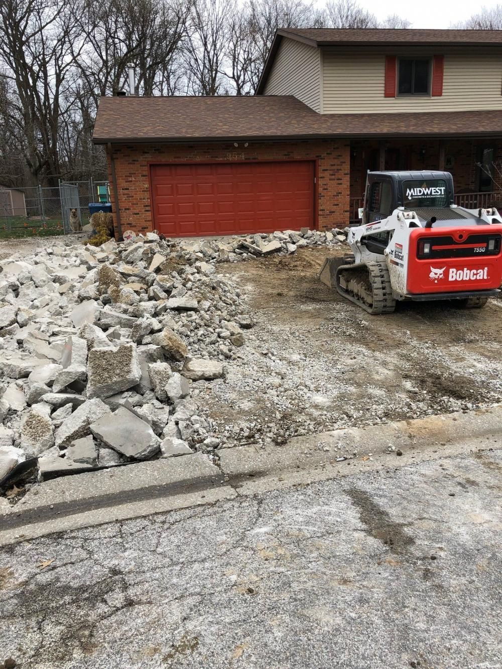 Bobcat skid steer clearing rubble from a driveway beside a brick house and red garage door.