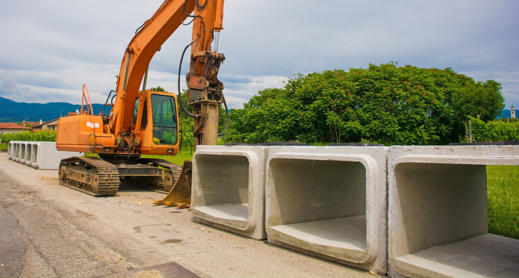 Excavator at roadside construction site beside large concrete drainage culverts under cloudy sky