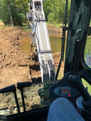 Excavator digging muddy trench beside a wooded pond, viewed from inside the cab