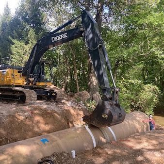 Excavator lifting a large concrete pipe in a wooded construction site with a worker nearby