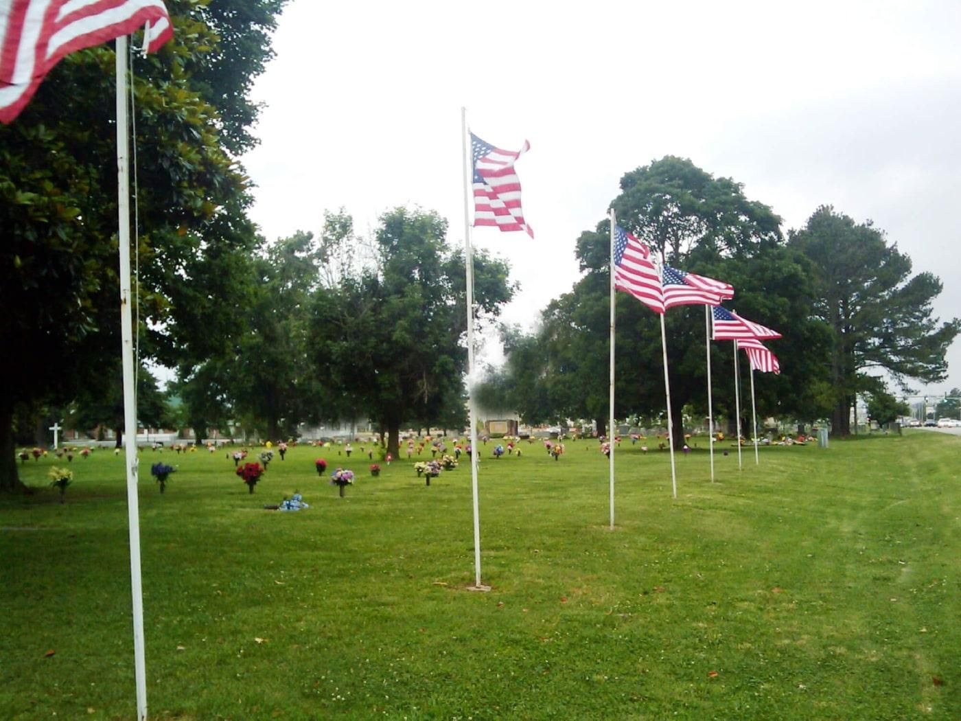 American flags on poles in a grassy cemetery. Trees and headstones fill the background.