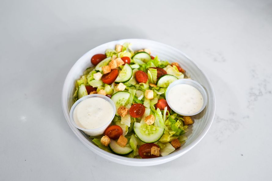 Bowl of mixed salad with cucumbers, tomatoes, lettuce, and two dressing cups