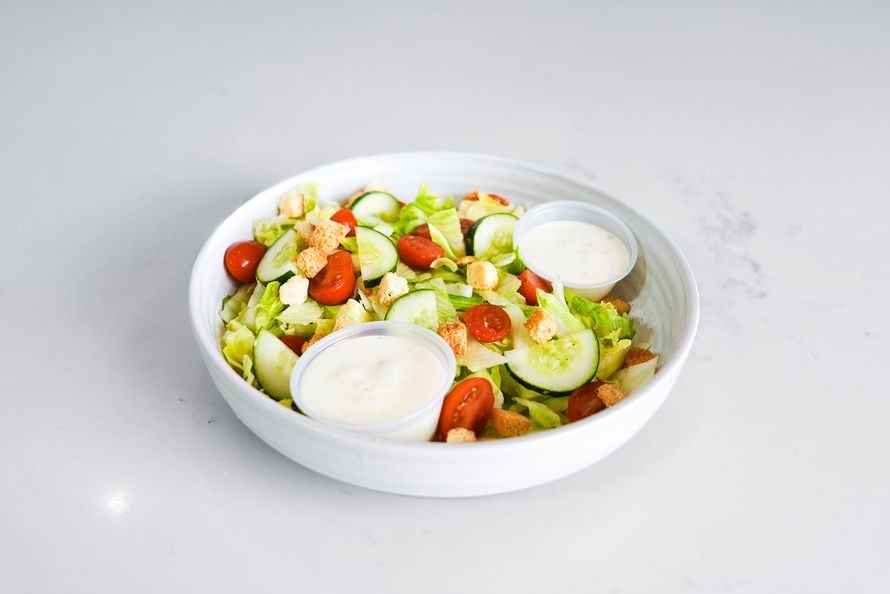 White bowl of salad with cucumbers, cherry tomatoes, croutons, and two dollops of dressing on a light background