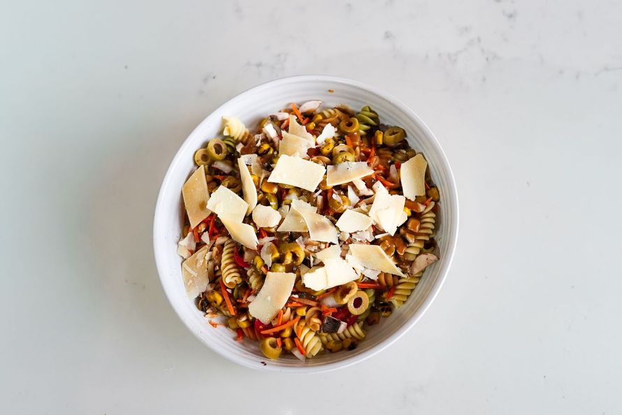 Bowl of pasta salad with cheese shavings and chopped vegetables on a white table