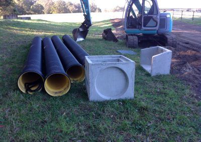 A pile of pipes and a concrete box are sitting on the grass next to an excavator.
