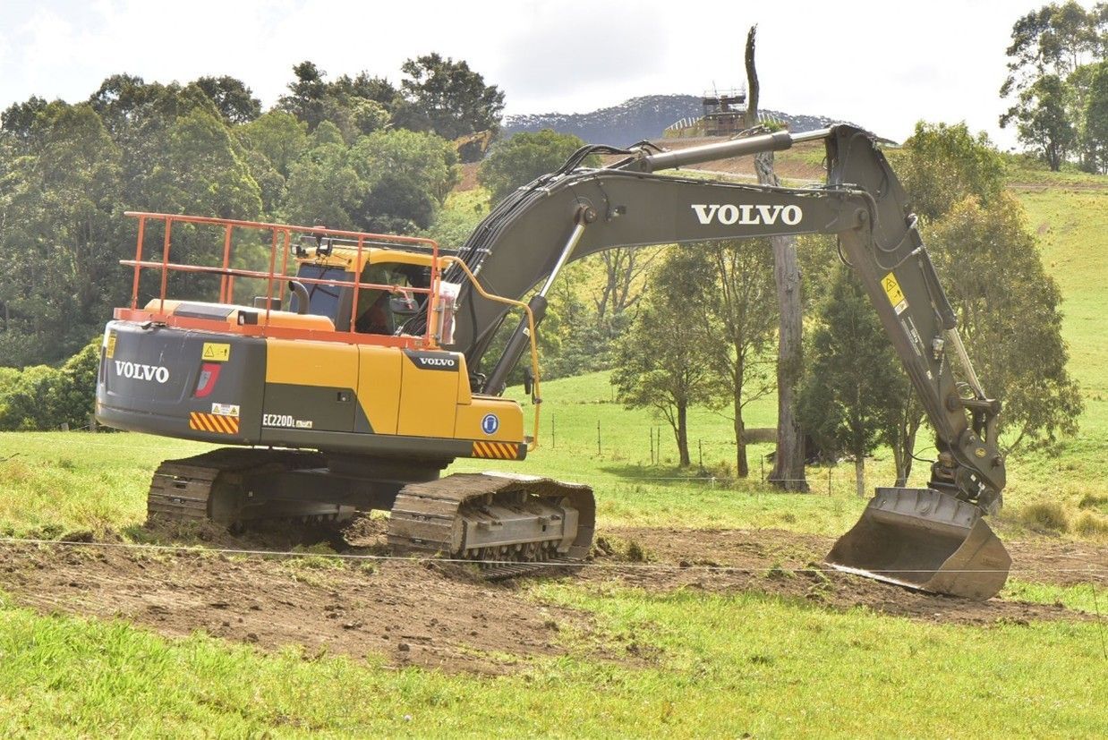 A yellow and black Volvo excavator digging in a grassy field with trees and hills in the background.