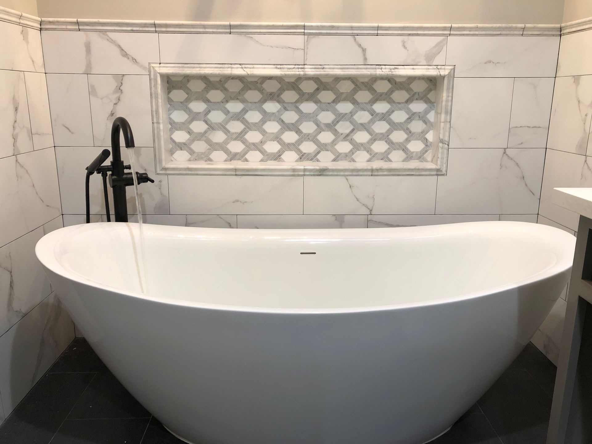 Freestanding white bathtub in a tiled bathroom with a decorative niche and black fixtures.