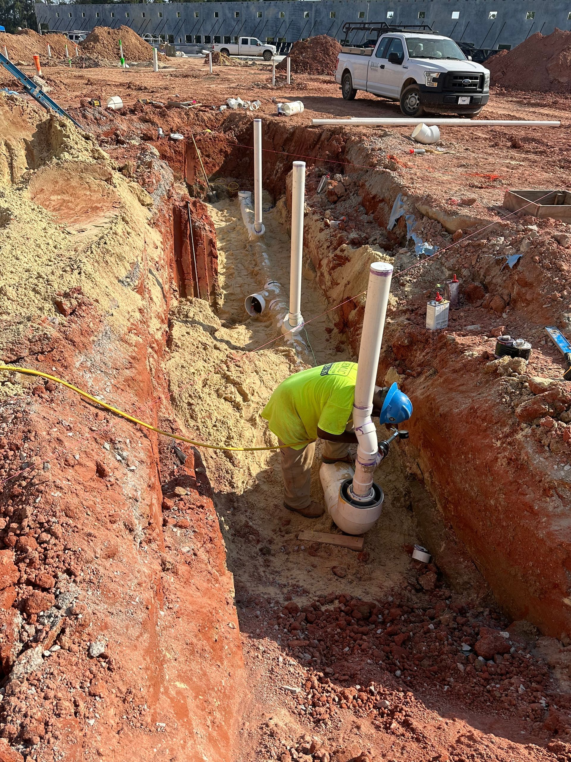 Construction worker in a trench connecting white pipes. Red dirt and a white truck are visible.