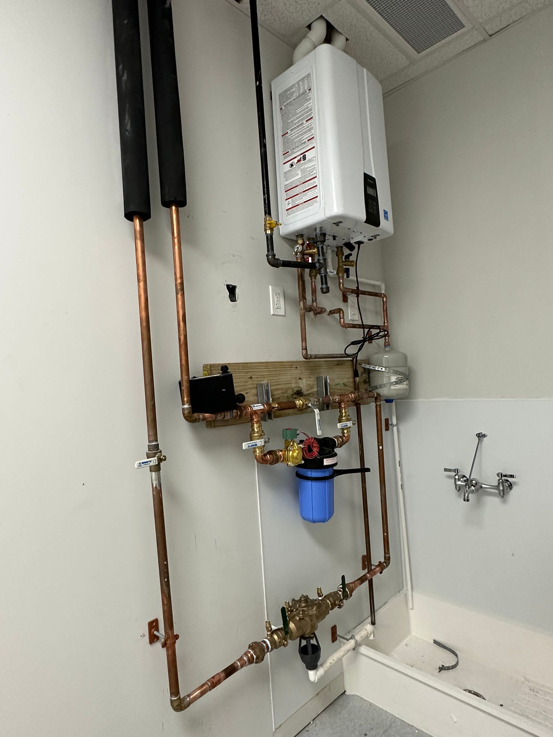 Copper pipes and a white tankless water heater mounted on a white wall. Blue water filter and valves are also visible.