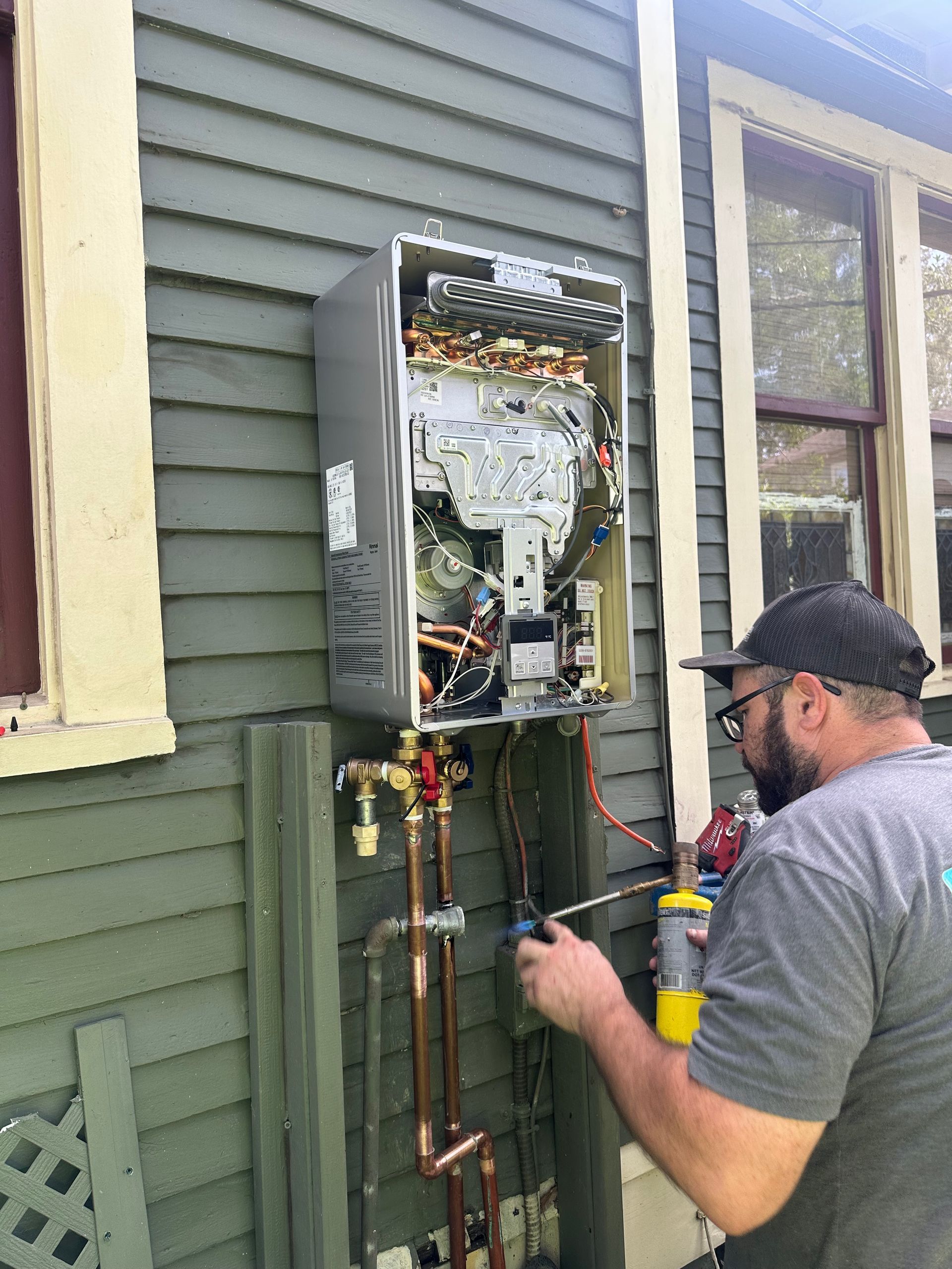 A plumber uses a torch to solder copper pipes near an outdoor water heater mounted on a green house.