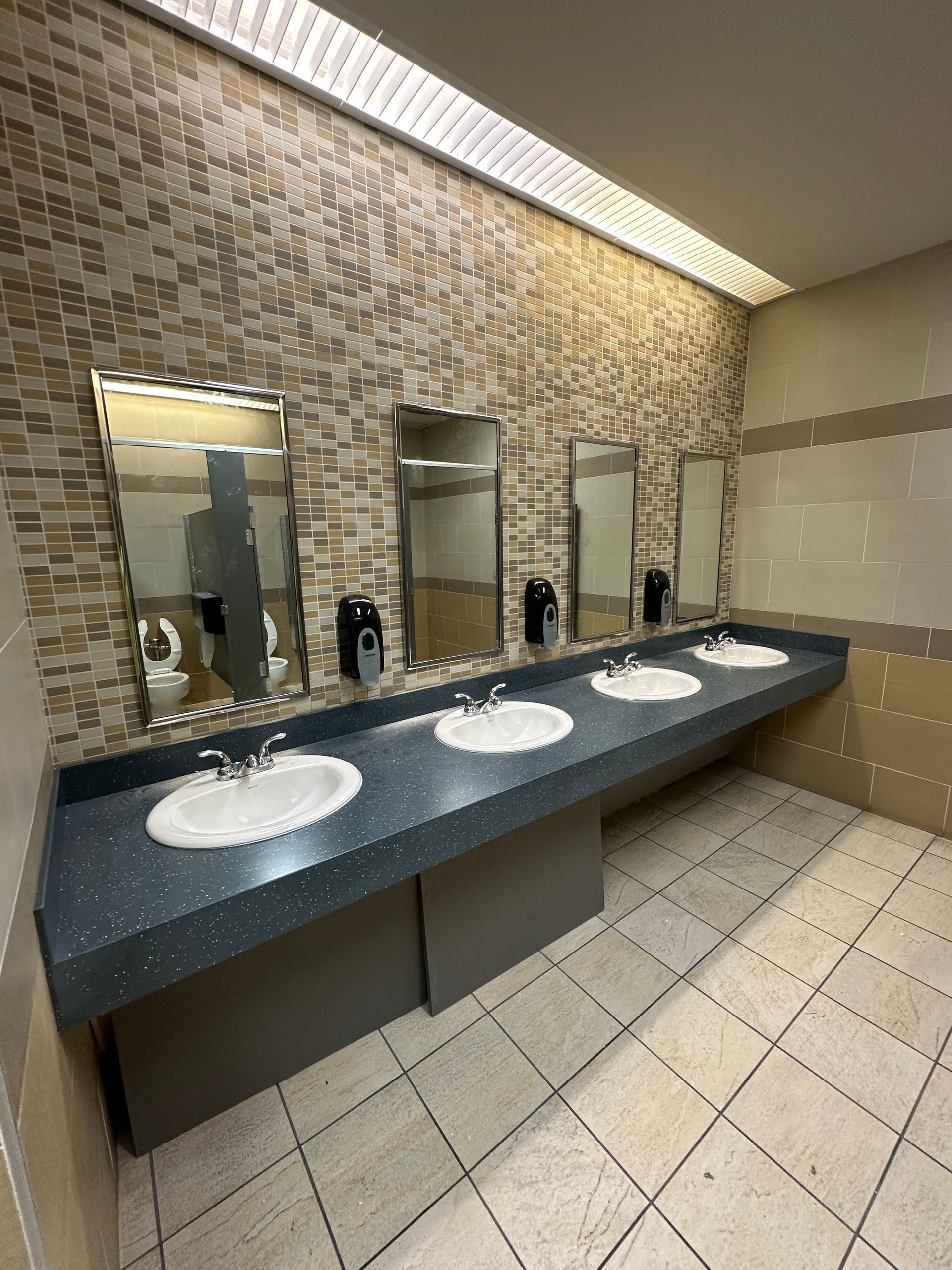 Public restroom with sinks, mirrors, and soap dispensers. Beige and brown tile walls, white sinks, and speckled countertop.