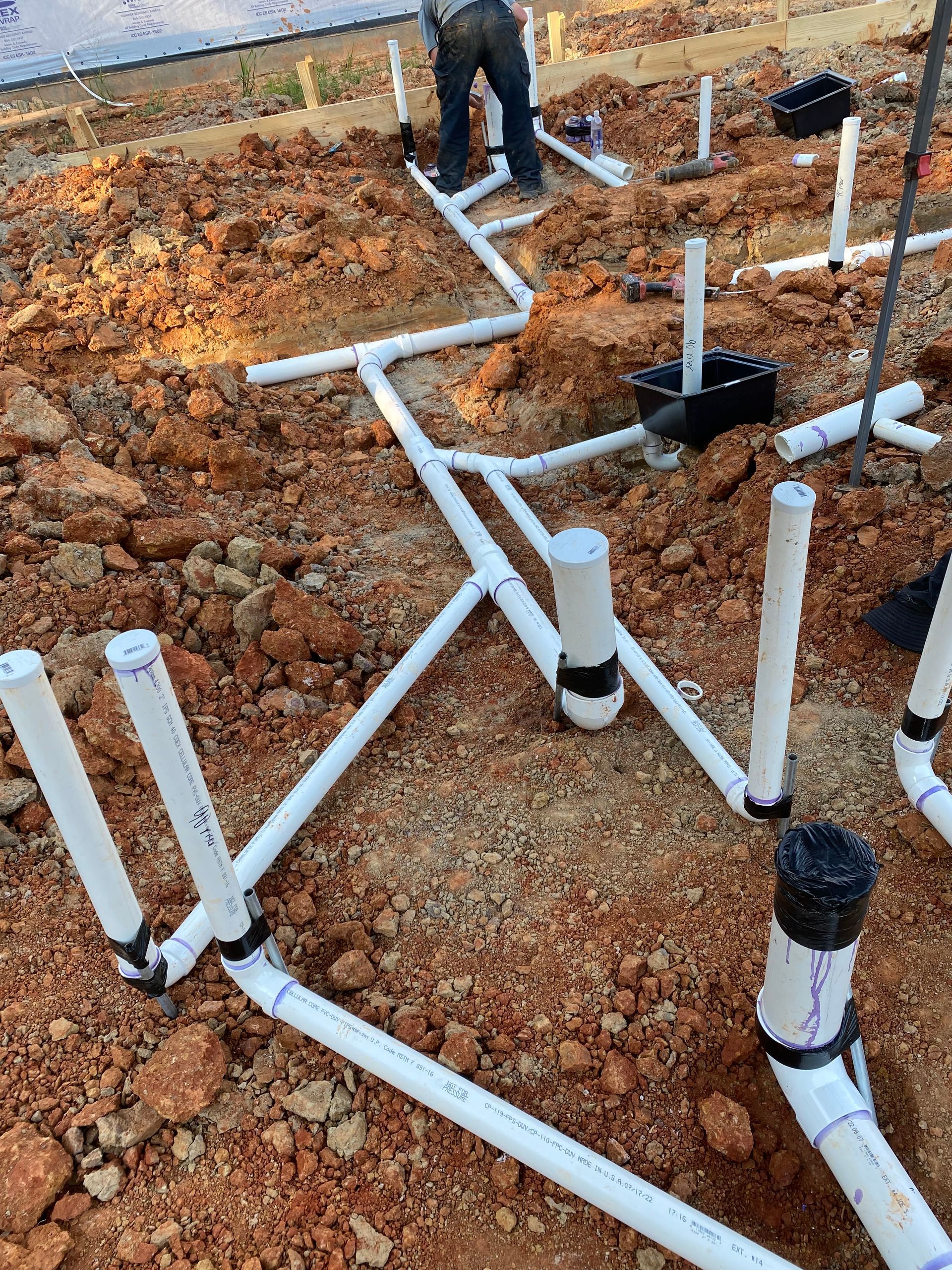 Plumber installing white PVC pipes in dirt for a building's drainage system.