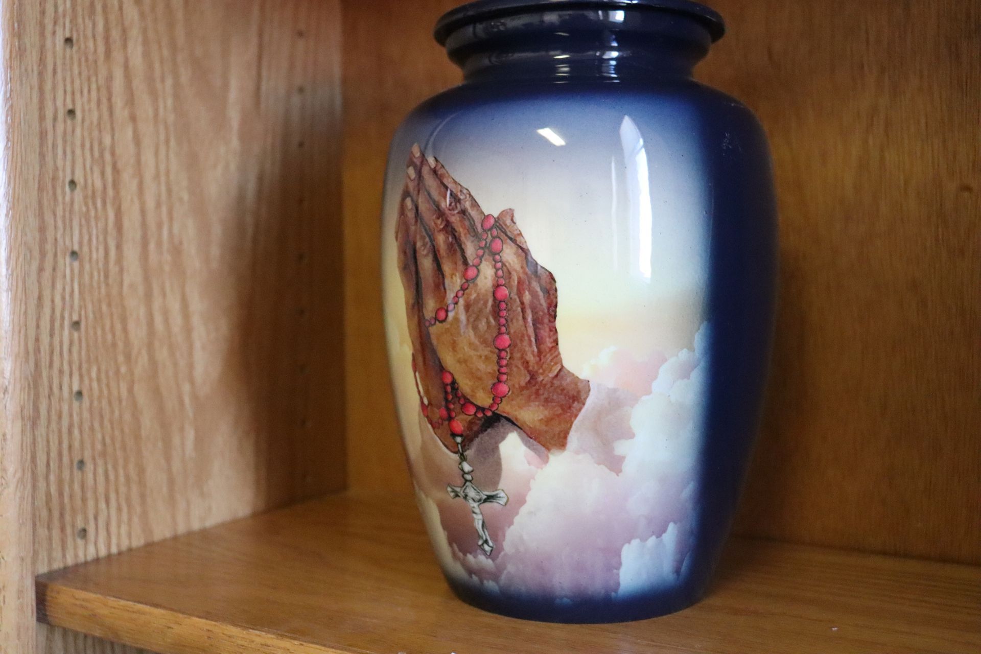 A blue vase with praying hands on it is sitting on a wooden shelf.