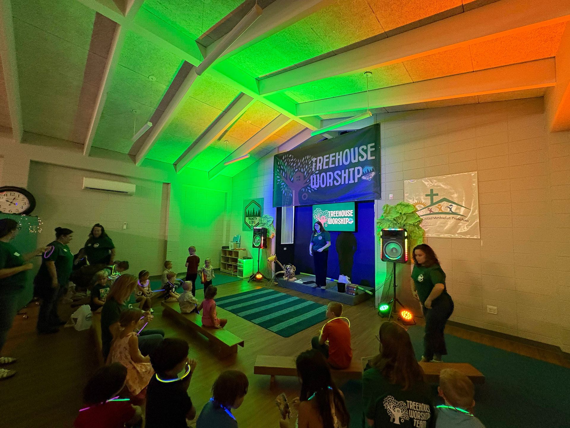 Indoor stage event with green lighting, audience seated on the floor, and a banner above the entrance