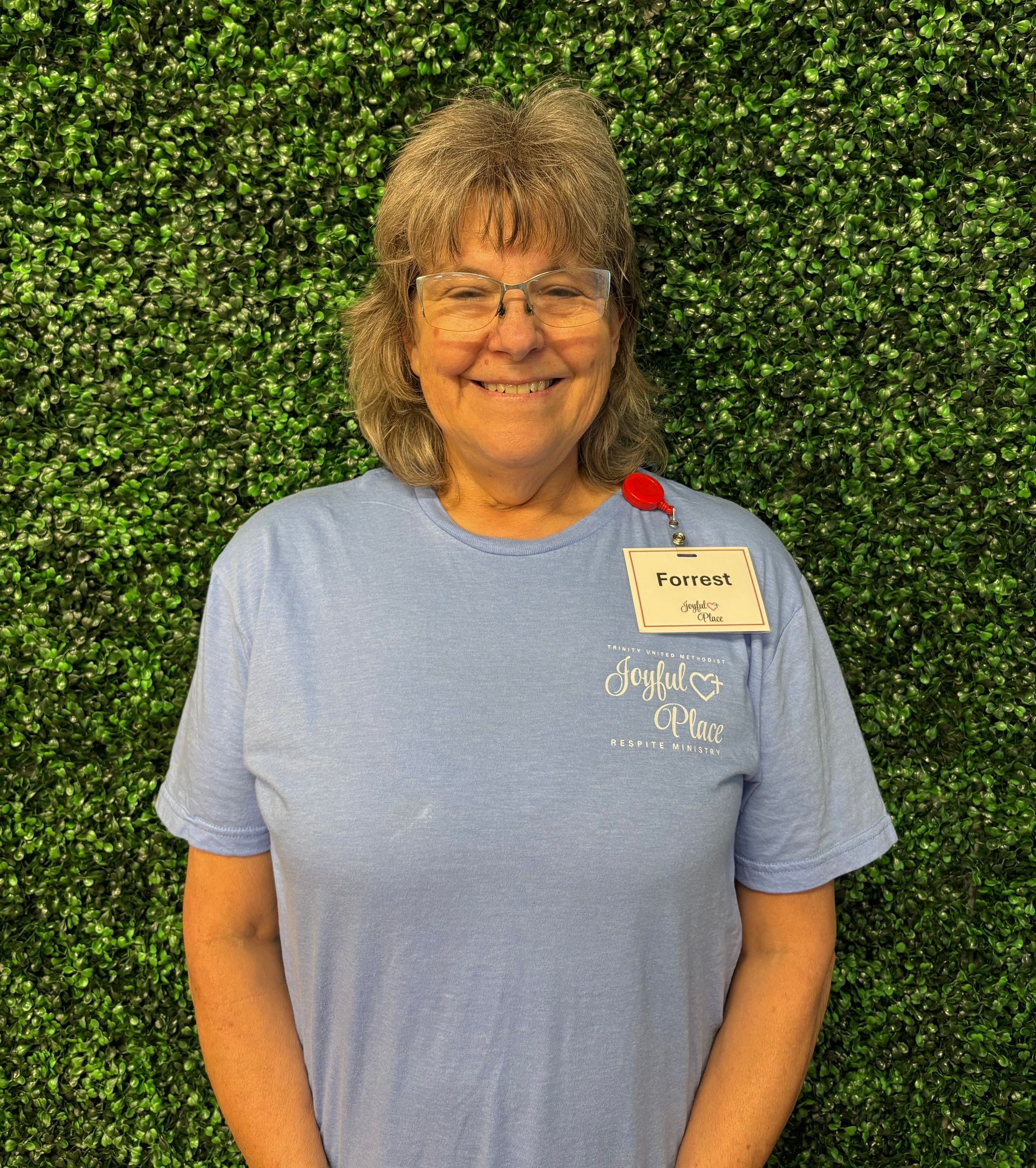 Smiling person in a light blue shirt with a name tag, standing before a green hedge wall.