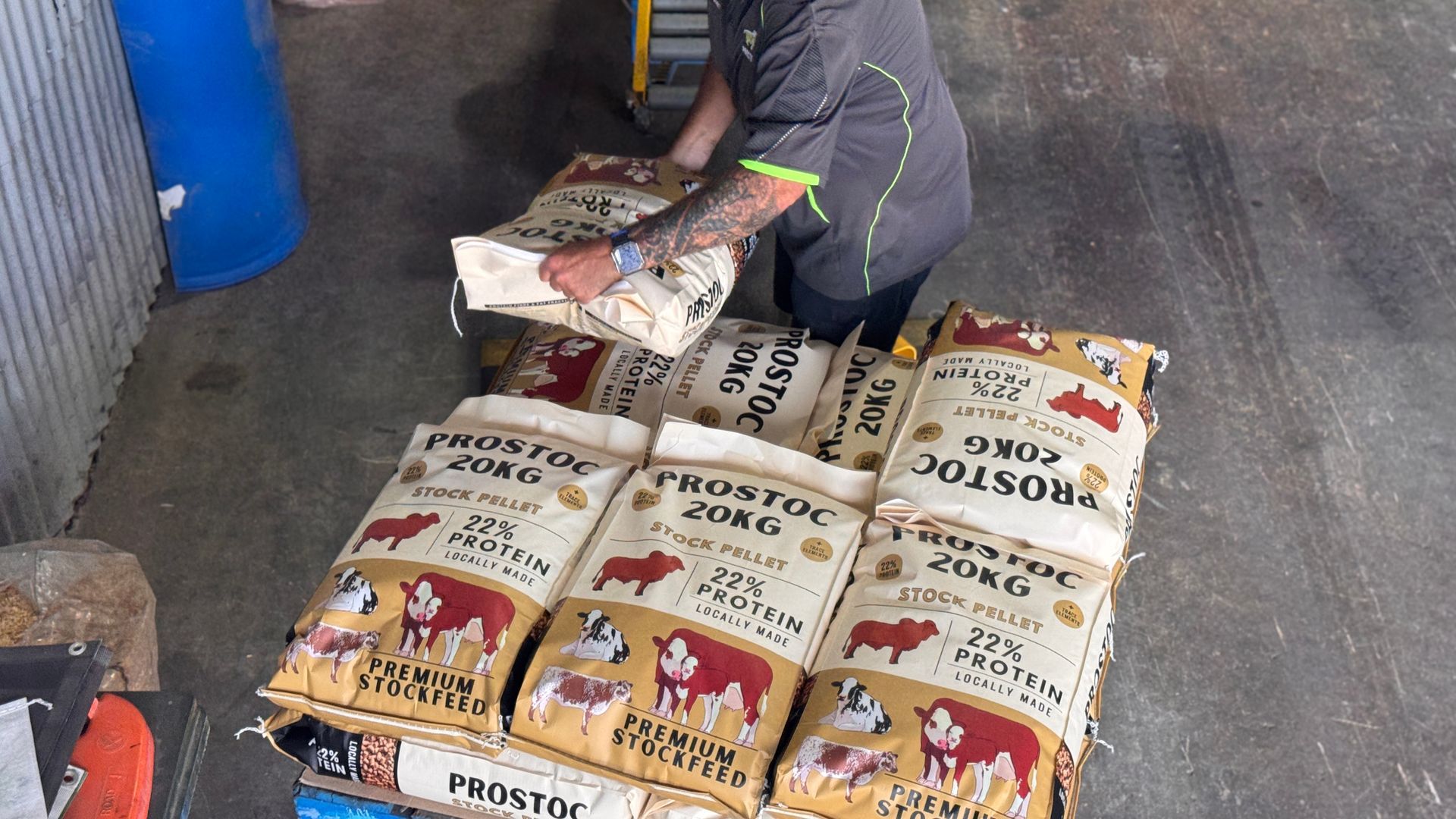 A person in a gray shirt stacks bags of Prostoc 20kg feed onto a pallet in an industrial setting.