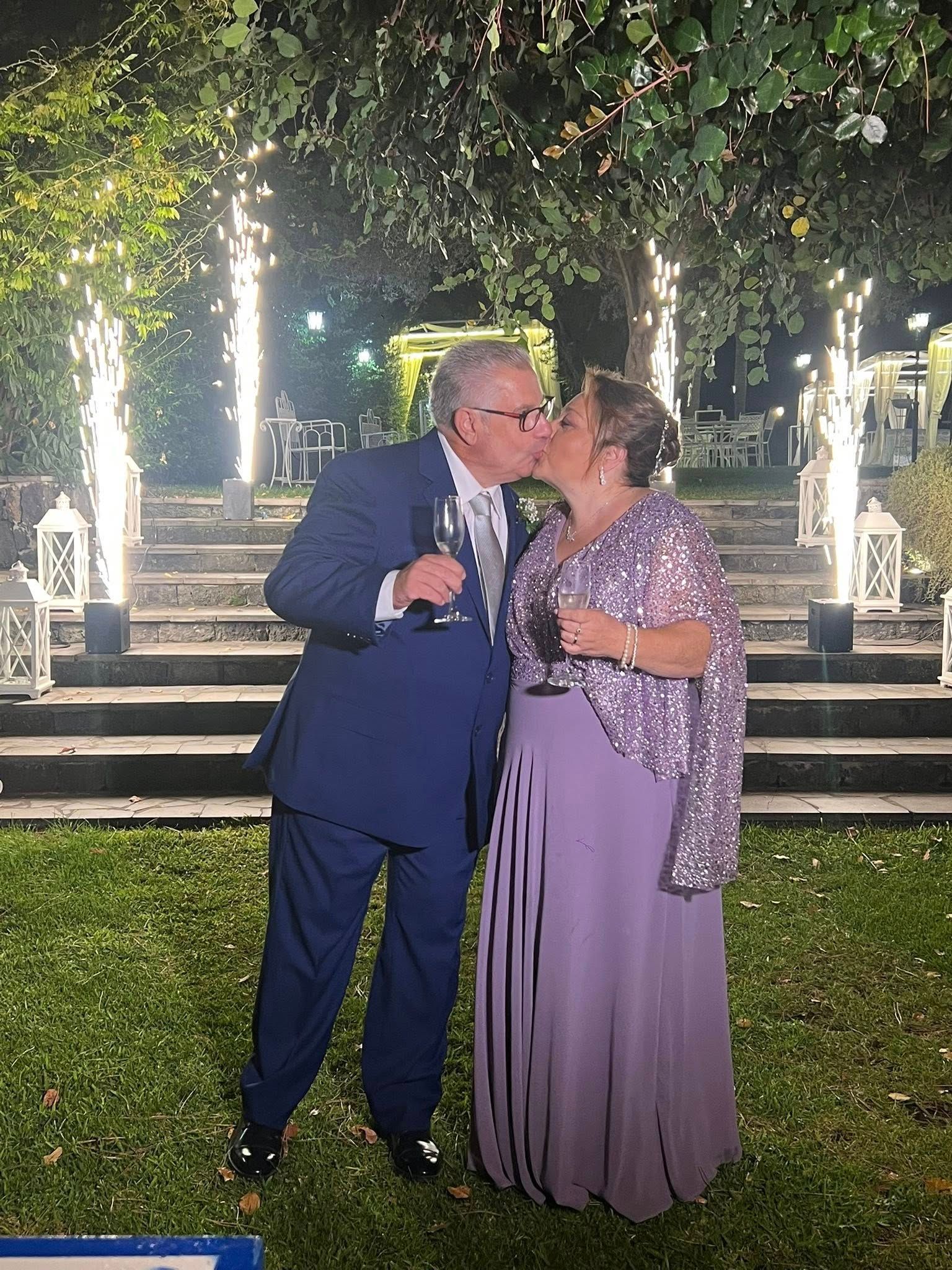 Coppia che si bacia all'aperto, festeggiando. Uomo in abito blu, donna in abito viola e scialle. Scintille e scale sullo sfondo.
