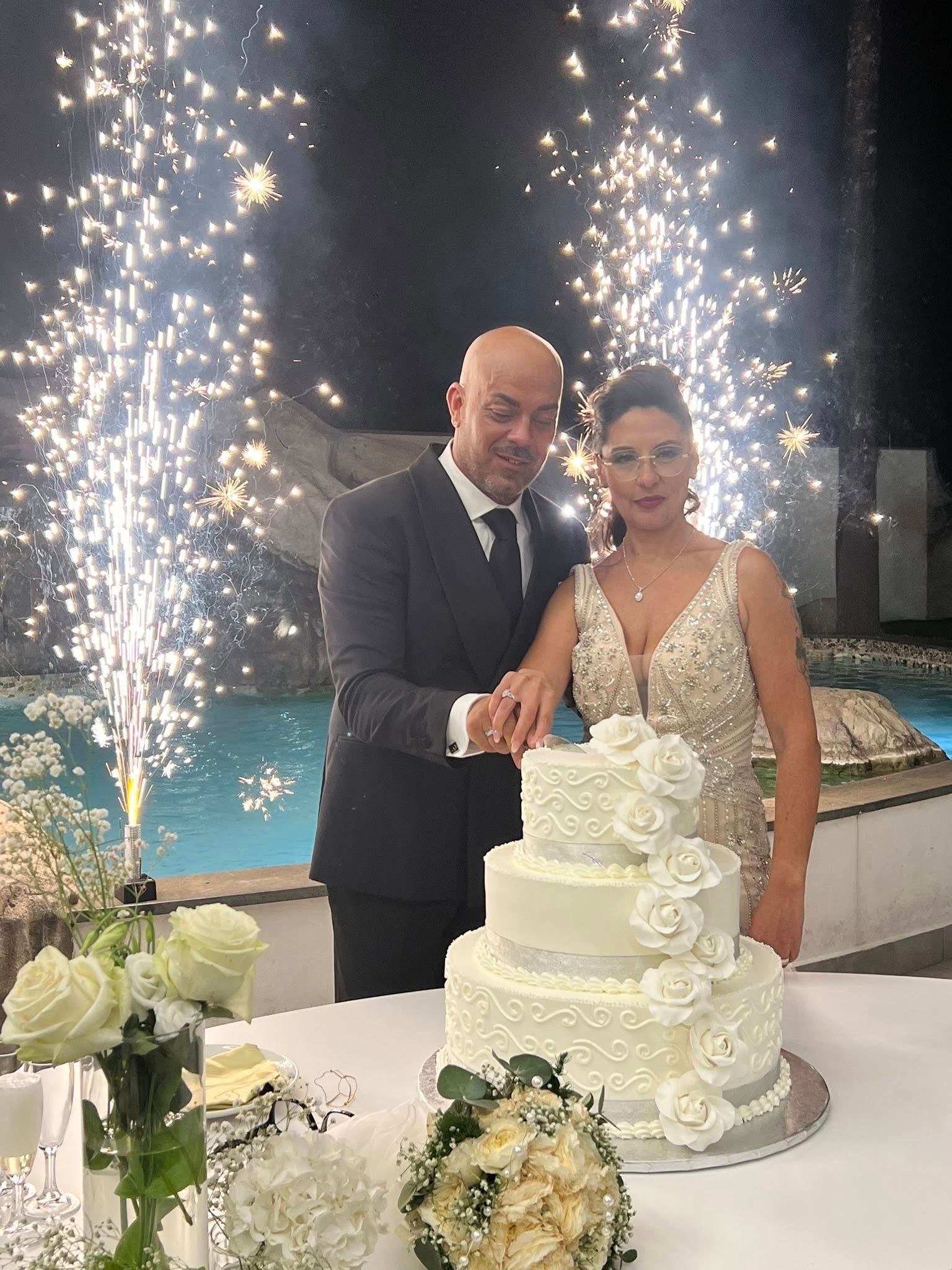 Una coppia taglia la torta nuziale con fuochi d'artificio sullo sfondo, vicino a una piscina.