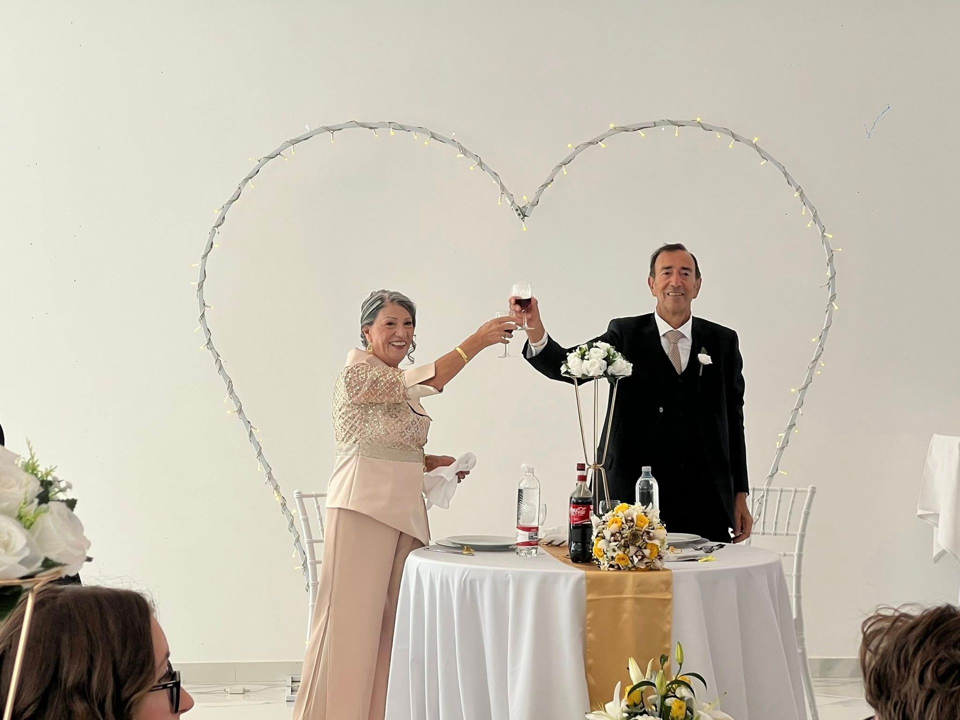 Una coppia brinda con un drink durante un ricevimento di nozze, davanti a una mostra di luci a forma di cuore.