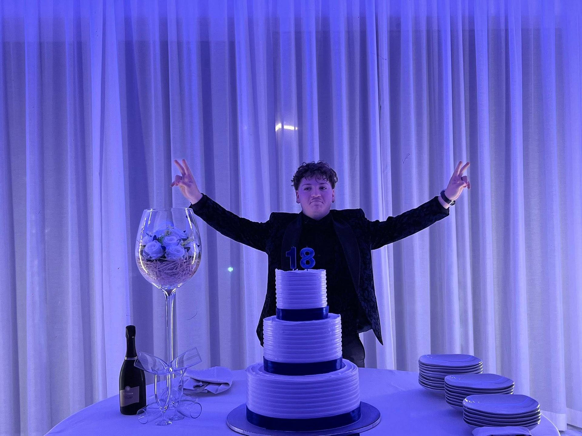 Giovane a una festa con torta, braccia alzate, festeggia. Sfondo con drappeggi bianchi, luci blu.