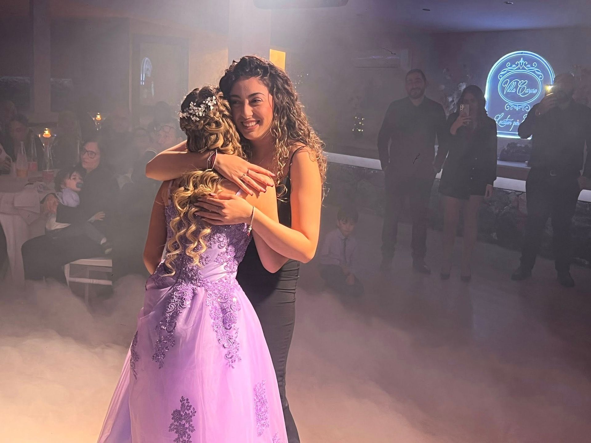 Una donna abbraccia un'altra che indossa un abito lilla su una pista da ballo con la nebbia.