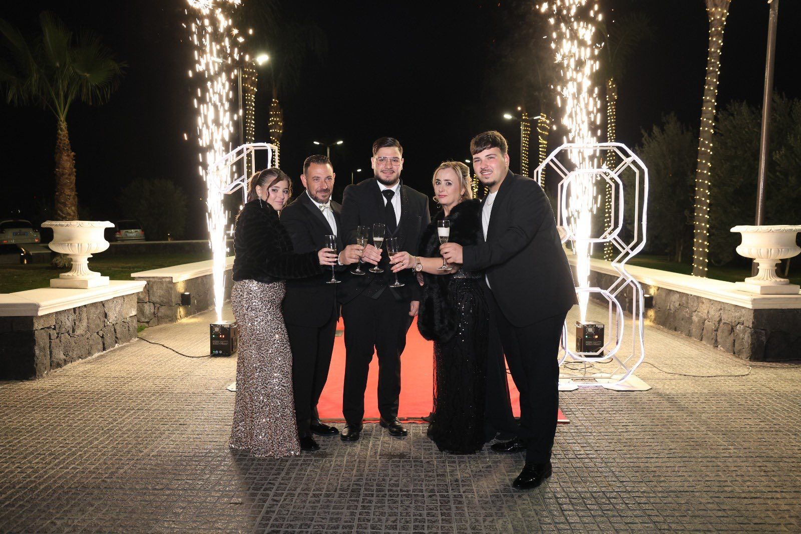 Un gruppo in abiti formali brinda su un tappeto rosso con fuochi d'artificio e il numero 8 illuminato sullo sfondo.